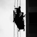 Two workers silhouetted on a suspended scaffold against a building facade.